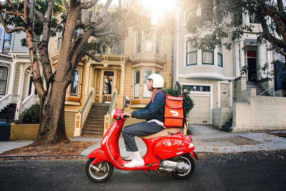A Doordash driver on a red scooter riding through a suburban street.